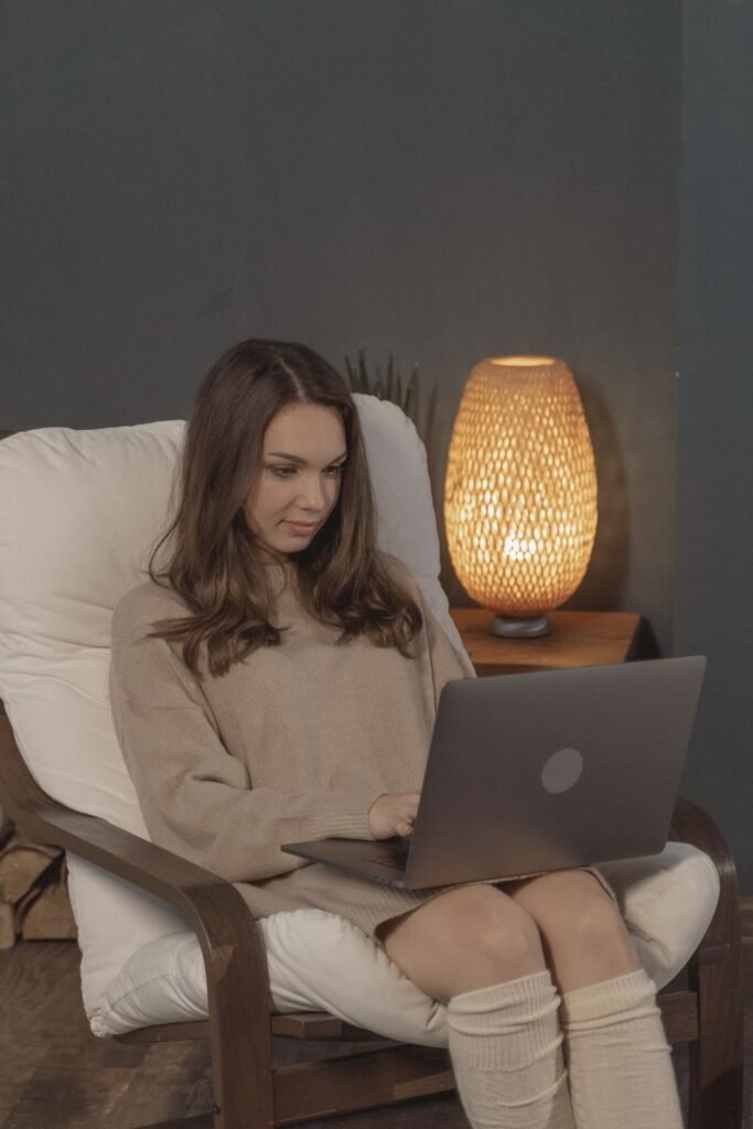 Woman relaxing in a cream armchair with a laptop on her lap, next to a glowing table lamp